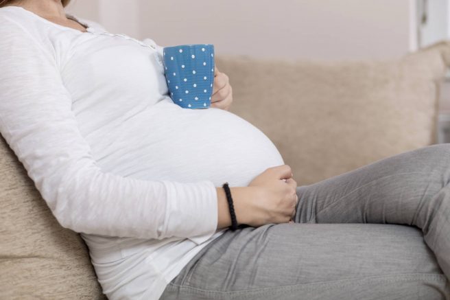 Femeie gravida stand pe o canapea cu o cana de ceai pe burta