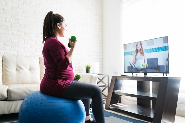 femeie-insarcinata-care face-exercitii-fizice-in-sarcina-stand-pe-o-bila-si-cu-greutati-in-maini-si-uitandu-se-la-televizor