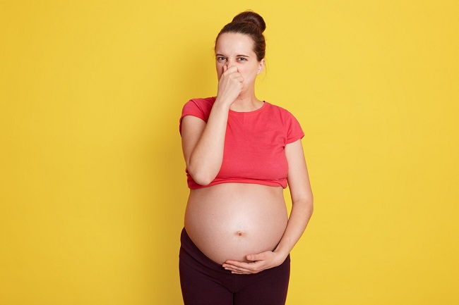 femeie-gravida-cu-burta-goal-care-se-ține-mâna-la-nas-de-la-gaze-în-sarcină-iar-în-spatele-ei-este-un-fundal-galben