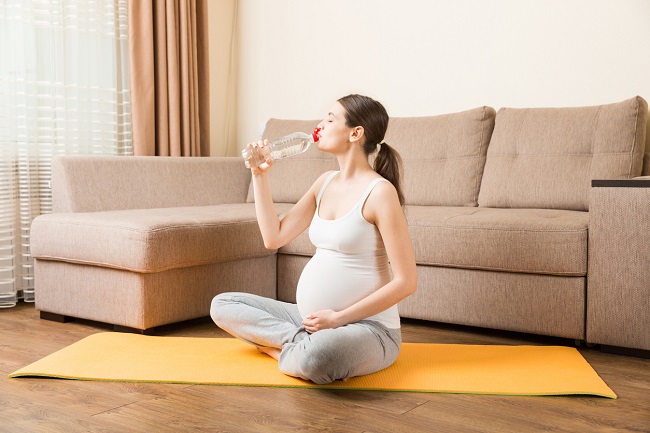 femeie-gravidă-așezată-pe-o-saltea-de-yoga-care-se-ține-cu-o-mână-de-burtă-și-bea-apă-în-același-timp