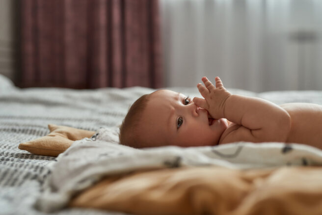 bebelus care sta pe pat cu degetul in gura

